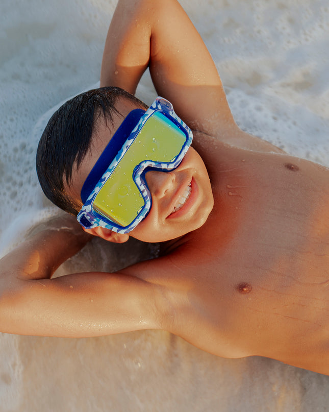 boy laying on beach in water wearing special ops kids' swim goggles in battleship blue showing a wide reflective lens and blue camo print