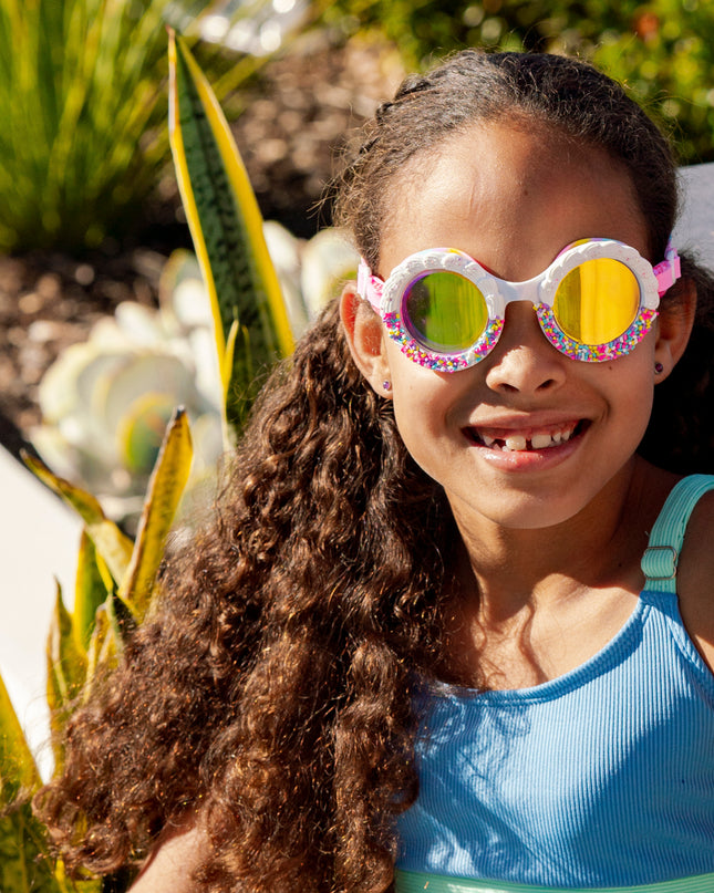 girl smiling poolside wearing bake off kids' round shaped swim goggles in color burst rainbow showing faux frosting detail and rainbow sprinkles