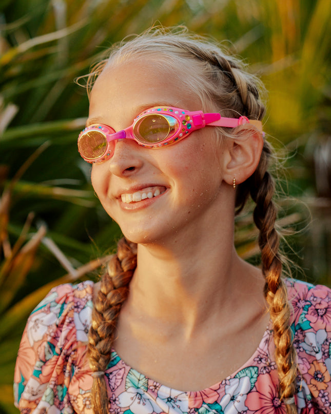 girl smiling poolside wearing glimmering gemstones kids' swim goggles in tropical tanzanite showing rhinestones and gems on a bright pink swim goggle frame