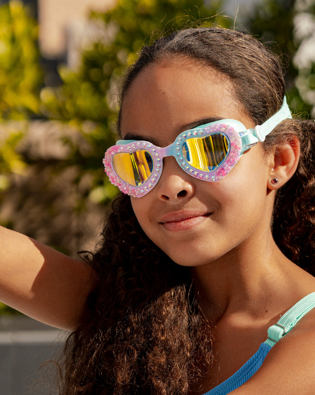 girl smiling outside wearing seaquin kids' heart shaped swim goggles in bluetiful showing details of crushed pink rhinestones and a set of rhinestones 
