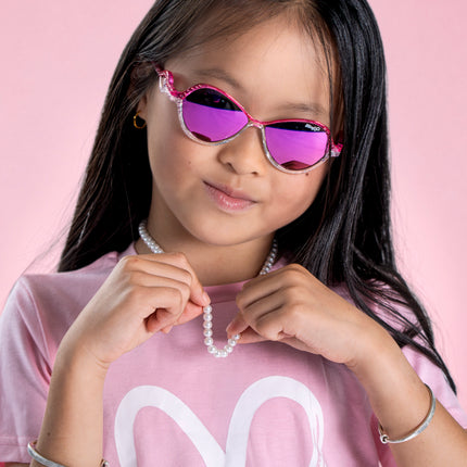 girl posing in pink studio wearing heart beach kids' sunglasses in color rosa romance featuring oval lens with a pink and white glittering frame and reflective lens