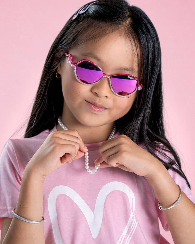 girl posing in pink studio wearing heart beach kids' sunglasses in color rosa romance featuring oval lens with a pink and white glittering frame and reflective lens