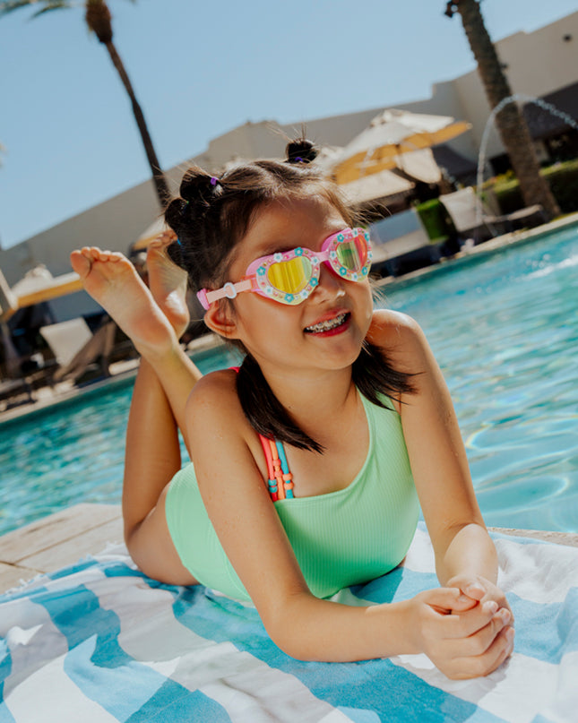 lifestyle of girl laying on beach towel poolside wearing daisies at dawn heart shaped swim goggles