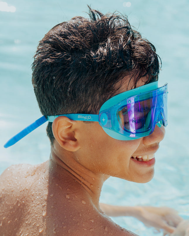 lifestyle of boy smiling sitting in pool wearing hologram kids swim goggles in polar stream blue showing frameless lens and blue colored silicone