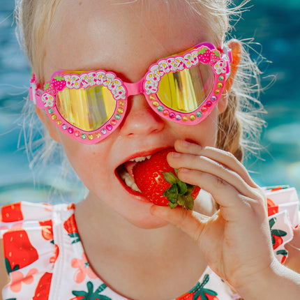 kid biting into strawberry wearing strawberry swimsuit and shortcake kids swim goggles in strawberry cream showing strawberry shortcake detail and rhinestones on the frame