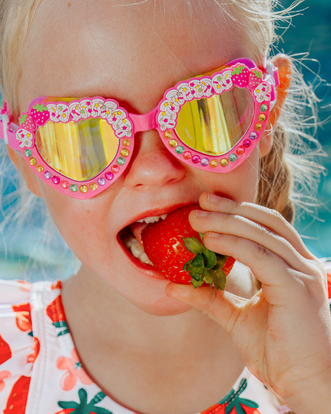 kid biting into strawberry wearing strawberry swimsuit and shortcake kids swim goggles in strawberry cream showing strawberry shortcake detail and rhinestones on the frame