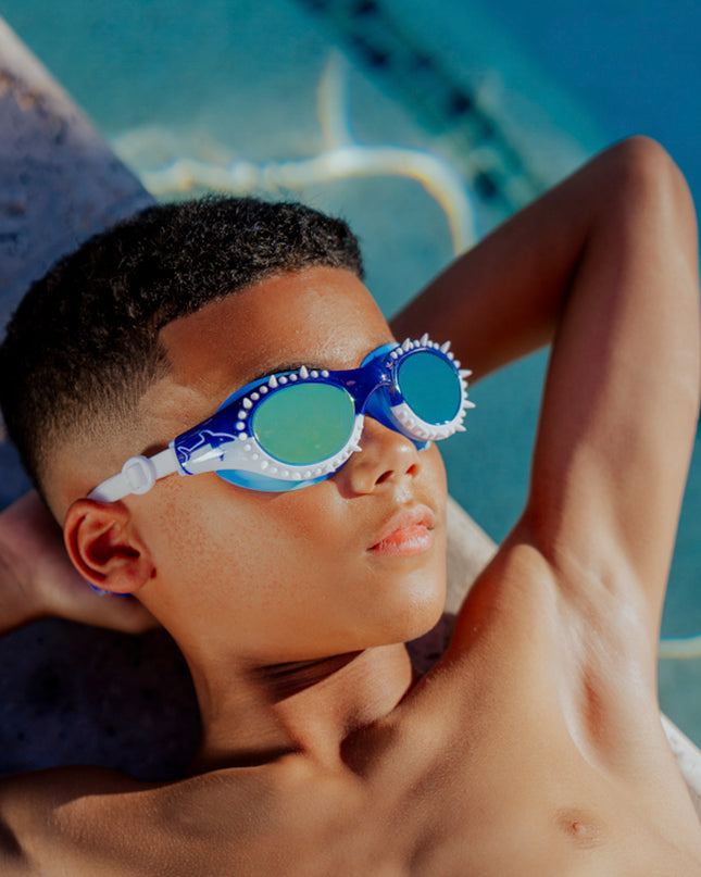 boy laying next to pool relaxing wearing shark surge kids swim goggles in great white strike showing silicone spike detail and shark printed frame