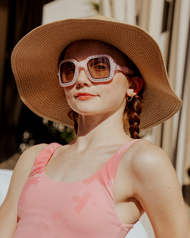 teen girl sitting poolside wearing vibrancy swim goggles in rose quartz showing a pale pink large square frame, rhinestones and tined lens